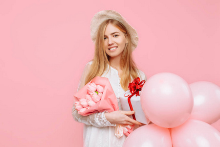 快乐的年轻女子穿着一件白色连衣裙，站着一束鲜花，一个礼品盒和粉红色背景上的气球。 三月八号概念