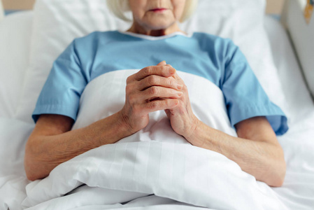 老年妇女双手交叉躺在床上，在医院祈祷