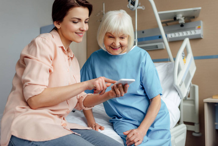 快乐的老年妇女和女儿坐在床上，在医院里使用智能手机