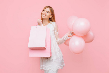 穿着白色连衣裙，手里拿着袋子，粉红色背景上有气球的快乐年轻女人