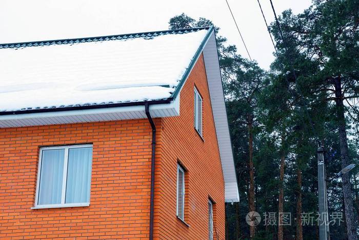 一座砖红色房子的一部分屋顶上有白雪背景是青松和天空