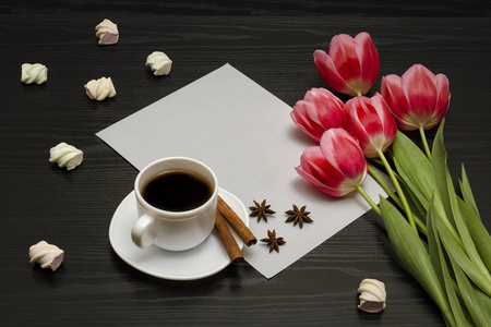 假日概念。一束粉红色的郁金香，一杯咖啡，棉花糖和一张黑色木制背景上的纸。