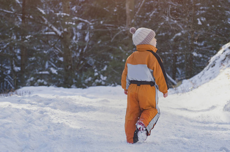 穿着橙色连衣裙的小男孩在一片针叶林的雪覆盖的道路上行走。 冬天阳光明媚的一天。 后景。