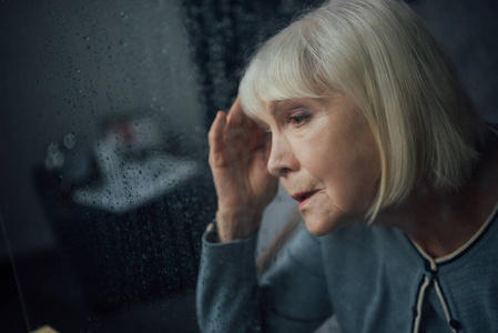 悲伤的老年妇女在家头痛的肖像，透过窗户和雨滴