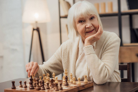 微笑的高级妇女坐在桌边，用手支撑下巴，在家下棋