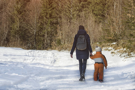母亲带着小儿子在针叶林的背景下沿着白雪覆盖的道路行走。 冬天。