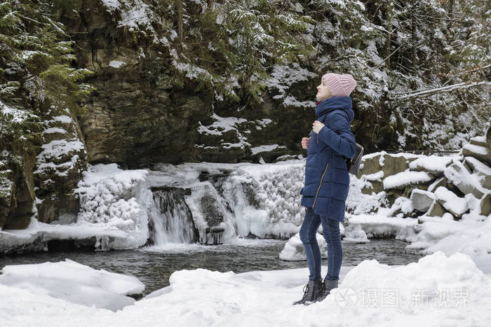 穿着蓝色外套的女孩站在冰雪覆盖的针叶林中的冰瀑布和岩石的背景上。 冬季阴天。 侧视。