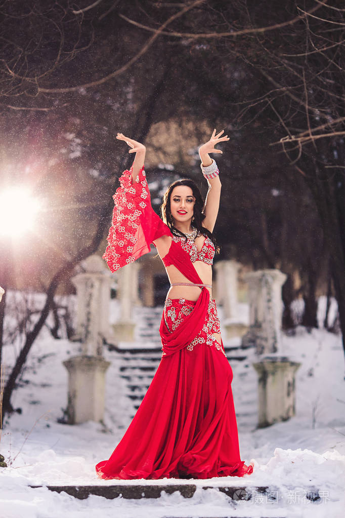 美丽的年轻女孩跳舞肚皮舞在冬季在雪上的公园红色礼服