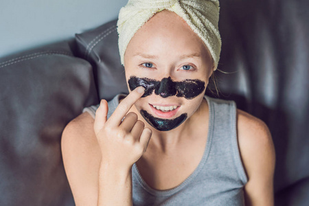 美丽的年轻女子在家里用面罩放松。愉快的快乐的妇女在面孔应用黑色面具