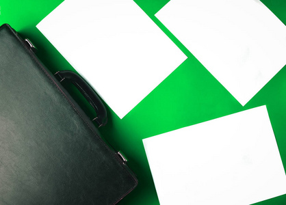 黑色皮箱和散落在绿色背景上的白色纸张。 复制空间