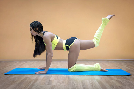 侧视年轻健身妇女做腿摆动为强壮和紧密的大腿。 有吸引力的运动女孩站在跪着的位置，在空的健身房大厅背景下在垫子上锻炼。