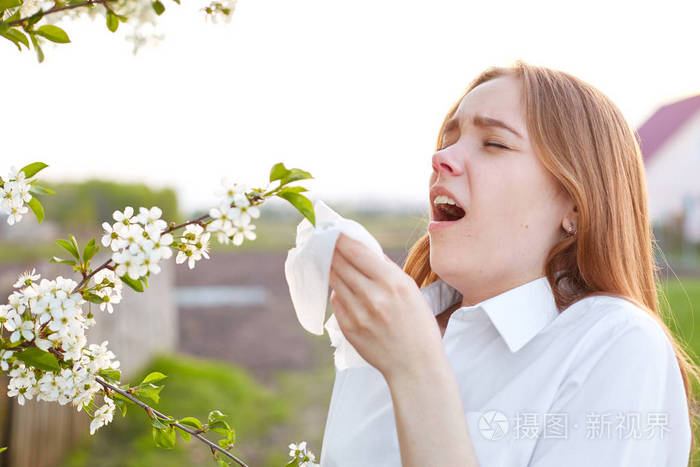 年轻女子总是用纸巾打喷嚏,春天站在附近开花,穿着优雅的衬衫,流鼻涕