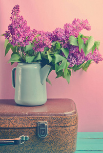 一束紫丁香在一个古老的漆漆茶壶上的老式手提箱在粉红色的背景。 复古风格仍然保留