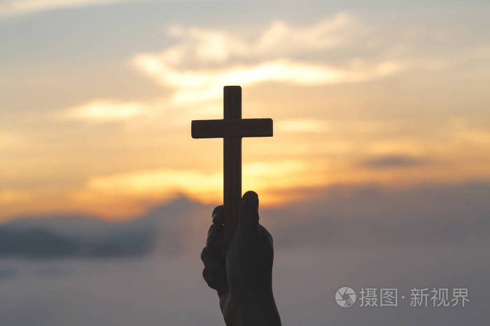 女性手的剪影,手持基督教十字架的圣举,背景是淡淡的日落.