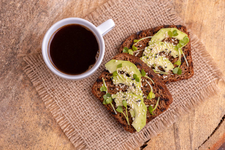 健康的黑麦烤面包与鳄梨和萝卜芽杯浓黑咖啡在Burlap餐巾纸。 木制背景。 简单的早餐。 顶部视图