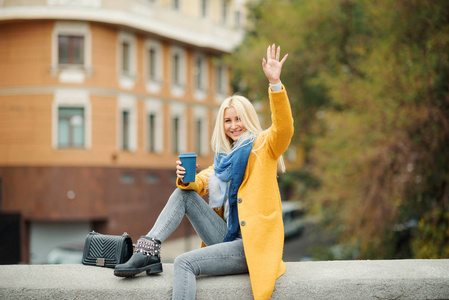 时尚快乐的年轻金发女人穿着明亮的黄色外套，拿着咖啡去。 走在街上。 一个年轻开朗的女人在工作休息时的肖像。 生活方式概念