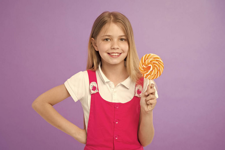 女孩微笑与棒糖在紫罗兰色背景。快乐的孩子与漩涡焦糖在紫色的背景。小孩子在棍子上微笑与糖果。食物和甜点。甜如棒糖。饮食和节食
