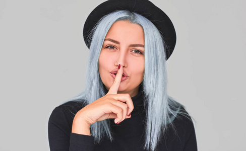 蓝色头发的肖像，女性穿黑色衣服，显示沉默标志，要求保密信息，在灰色工作室背景上有相当的表情。 秘密手势嘘。