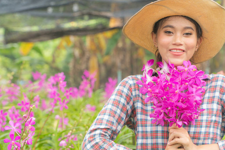 一位穿着格子衬衫戴着帽子的女园丁手里拿着一朵粉红色的兰花。 开心地笑着。 在兰花花园里