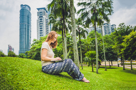 一个年轻的女人，穿便服在摩天大楼为背景的热带公园使用笔记本电脑。移动办公概念