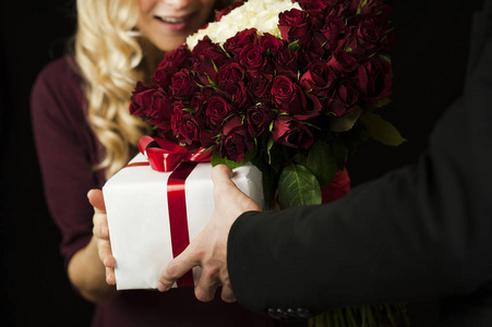 一个年轻人给一个礼物，一个白色的盒子，红色的蝴蝶结和花给一个女孩在一个孤立的黑色背景。情人节概念。情侣日。