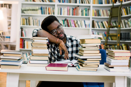 非洲裔美国人躺在桌子上，被图书馆里的书包围着。 学生又无聊又累。