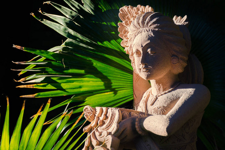 传统的巴厘岛砂岩女子欢迎祭品雕像在她的手中，阳光照亮了棕榈树。