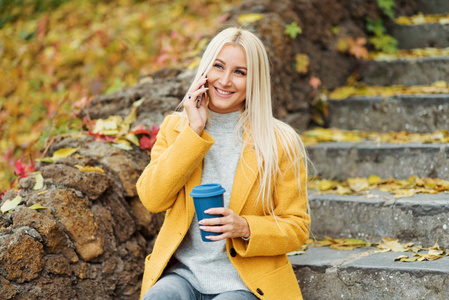 年轻的金发女人坐在城市公园里，喝着咖啡，用手机去