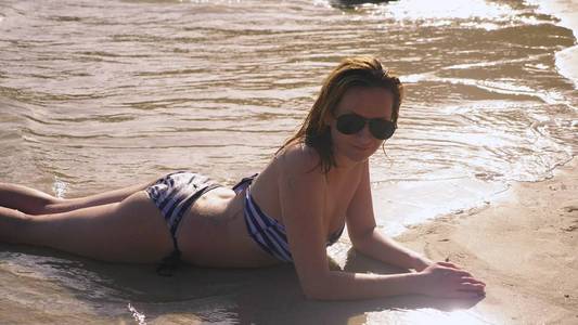 穿着游泳衣和黑色太阳镜的金发女孩在海边的白色沙滩上度假。美丽的模型与的身体游泳在蓝色的海水