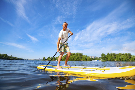 快乐的人在阳光明媚的日子在一条大河上的一块木板上划船。 站起来，桨，登机，令人敬畏的，积极的娱乐在大自然中。 大角度。