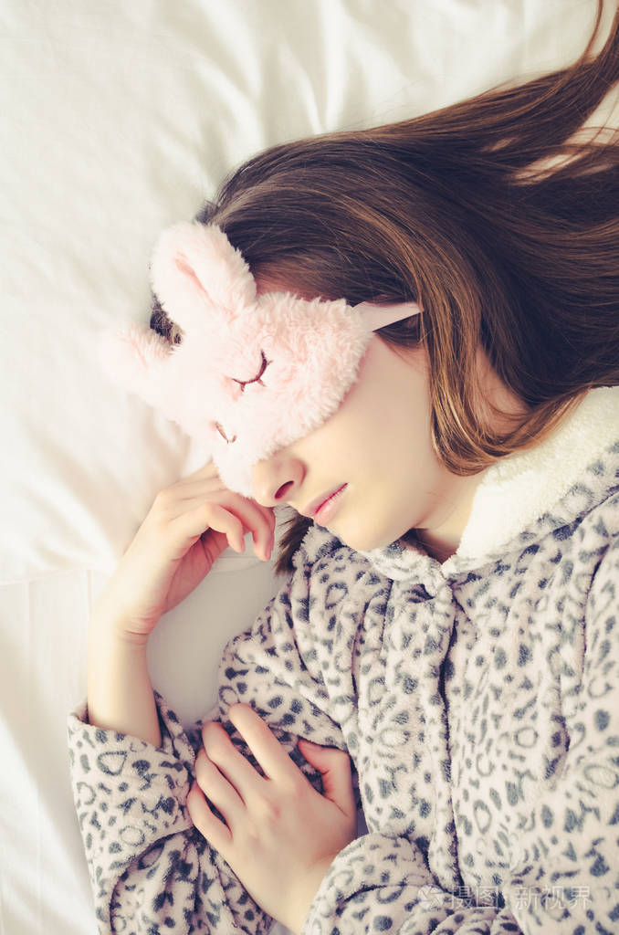 一个戴着可爱睡眠面具,穿着暖和睡衣的年轻漂亮女人躺在床上,早上睡觉