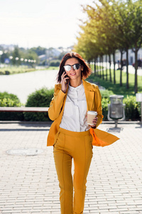 漂亮的女商人穿着黄色西装走在街上，喝咖啡，通过智能手机交谈。 智能设备概念商业生活方式沟通人。