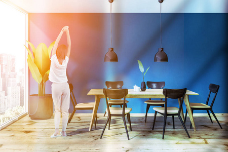 妇女在阁楼餐厅内部，蓝色墙壁，木制地板，木制桌子，黑色和米色椅子和两个时尚的天花板灯以上。 调色图像