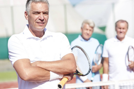 在阳光明媚的日子里，自信成熟的男子手持网球拍，双臂交叉站立在球场上对抗朋友