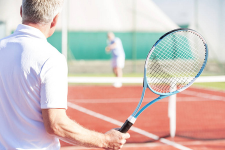 在网球场上与朋友玩耍时，成熟男人拿着球拍的后视图