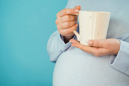 孕妇拿着陶瓷杯茶或咖啡在肚子上。穿蓝色衣服的年轻女孩穿着准妈妈。母亲母亲怀孕爱情概念