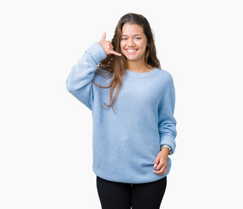年轻漂亮的黑发女人穿着蓝色的冬季毛衣，在孤立的背景上微笑，用手和手指做电话手势，就像在电话里说话一样。 交流概念。