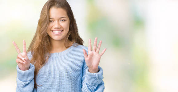 年轻漂亮的黑发女人穿着蓝色的冬季毛衣，在孤立的背景下，用手指举着七号，微笑着自信和快乐。