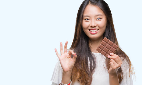 年轻的亚洲女性，在孤立的背景下吃巧克力棒，用手指做OK标志，很好的象征