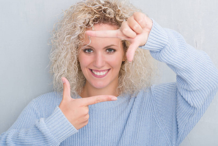 年轻的金发女人，卷曲的头发在灰色的背景上微笑，用手和手指制作框架，用快乐的脸。 创造力和摄影理念。