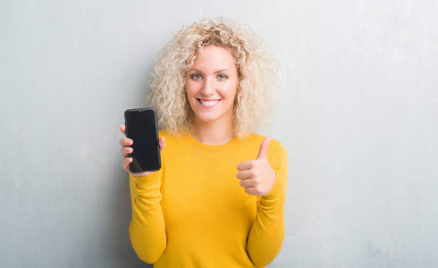 年轻的金发女人，在灰色的灰色背景上，显示着空白的智能手机屏幕，开心地大笑着，做着好的，用手指签署拇指，好的标志