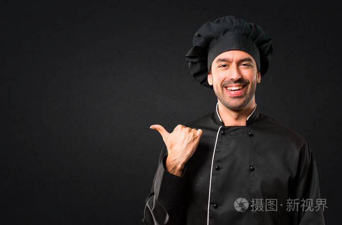 身穿黑色制服的厨师用手指指着旁边，一边展示产品或想法，一边期待着在黑色背景上微笑