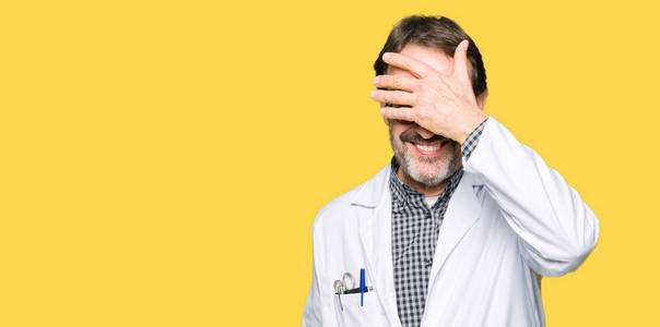 中年医生穿着医用外套的男人笑着，用手捂着眼睛，为惊喜。 盲目的概念。