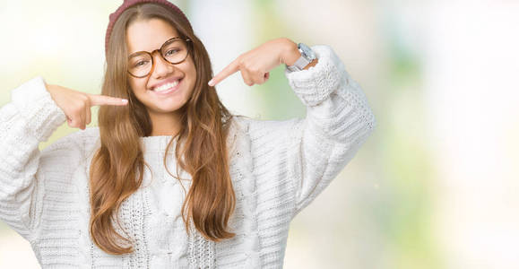 年轻漂亮的深色头发的女人戴着眼镜和冬天的帽子，在孤立的背景上微笑，自信地展示，用手指牙齿和嘴巴指着。 健康概念。