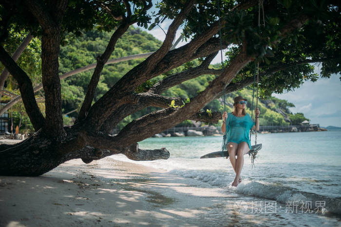 一个年轻的女人在海边一棵大树的树荫下荡秋千。 海滩上的女孩在热带岛屿的海岸摇摆。