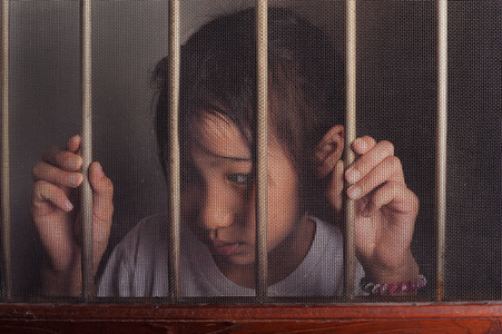 悲伤的亚洲孩子站在电线屏幕窗口后面。 不幸的孩子独自在家。 在黑暗的房间里心烦意乱