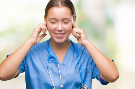 年轻的白种人医生女士穿着医疗制服，在孤立的背景上，用手指覆盖着耳朵，因为嘈杂的音乐声，她的表情很生气。 聋的概念。