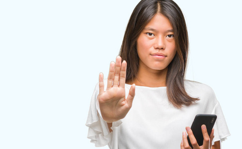 年轻的亚洲女人用智能手机在孤立的背景上发短信，张开手做停止标志，严肃自信的表情防御手势