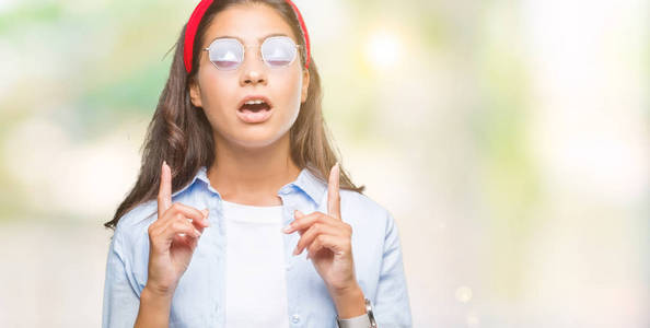 年轻漂亮的阿拉伯女人戴着太阳镜在孤立的背景上惊讶和惊讶地抬头，用手指和举起的手臂指着。