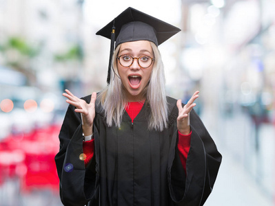 年轻的金发女人穿着研究生制服，在孤立的背景下庆祝疯狂和惊讶的成功，举起手臂和睁开眼睛尖叫兴奋。 赢家概念